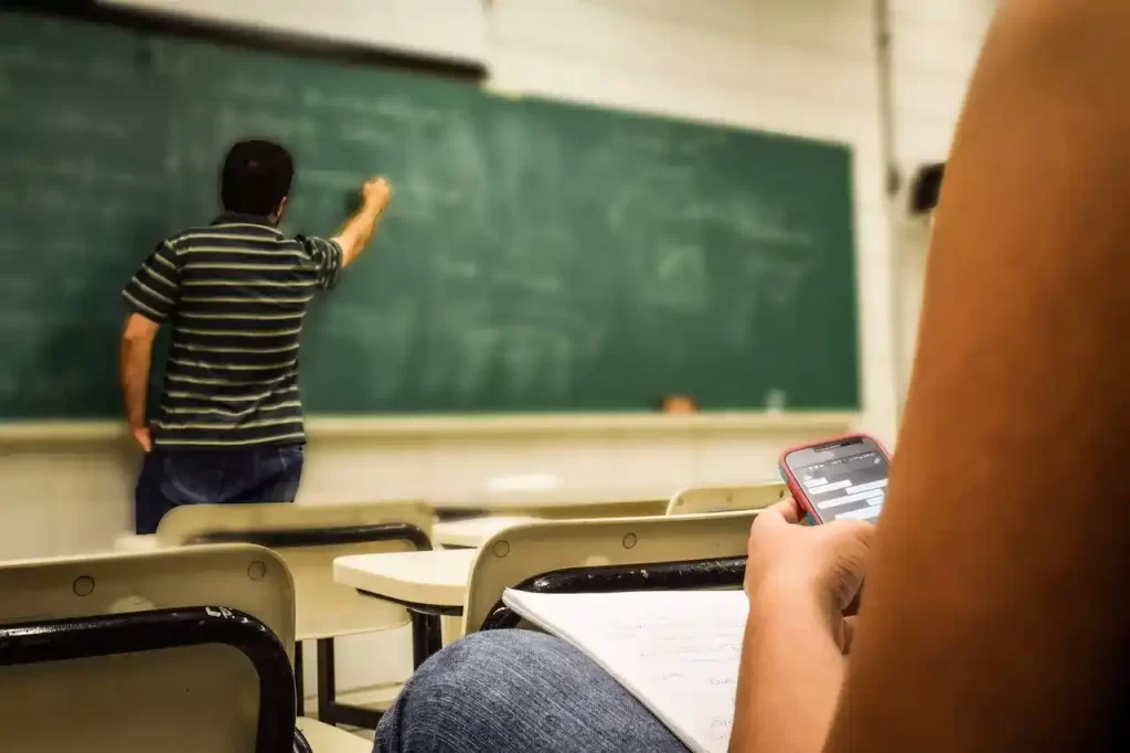 A student looking in their cellphone with the teacher is teaching in front of the class