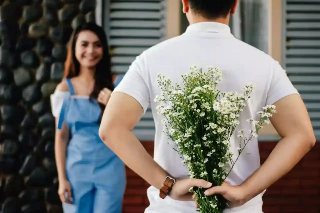 A guy holding a bouquet of flowers behind her while a girl is smiling at her