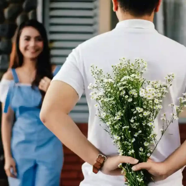 A guy holding a bouquet of flowers behind her while a girl is smiling at her