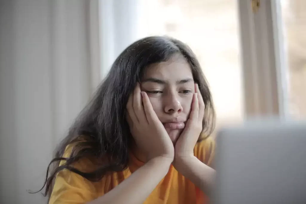 A woman sitting with her hands on her cheeks looking upset and tired at her computer screen