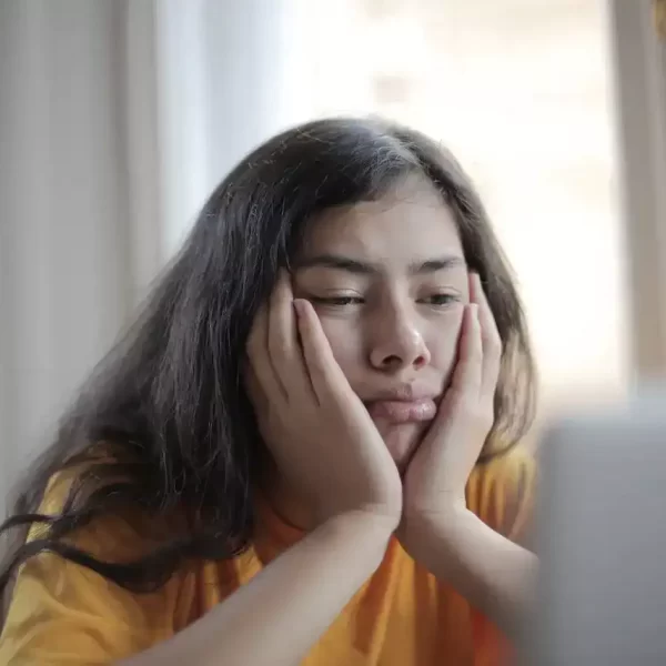 A woman sitting with her hands on her cheeks looking upset and tired at her computer screen