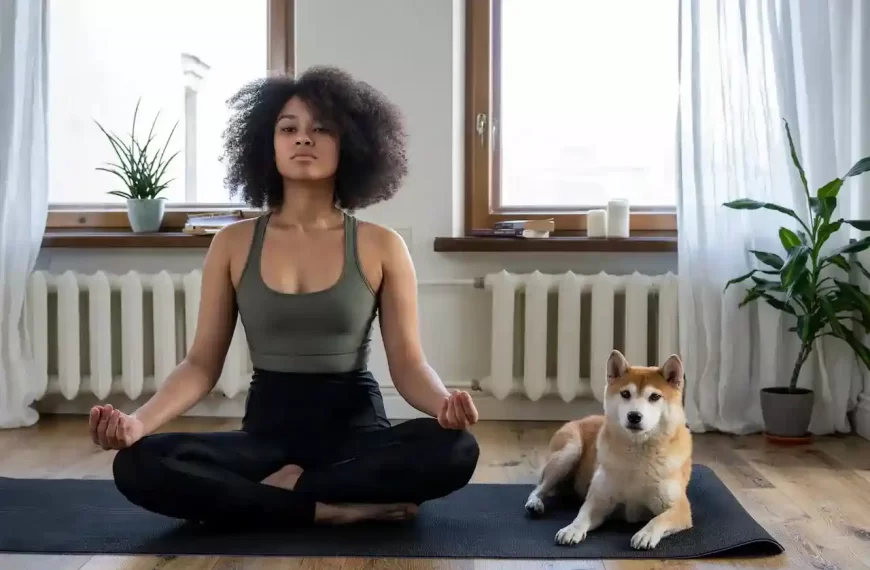 A woman meditating on a yoga mat with a dog is sitting besides her