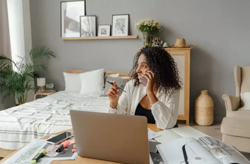 A woman sitting in front of a computer while talking on the phone, with a pen in her hand