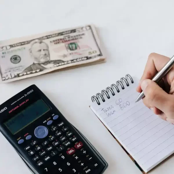 Close up of a persons hand writing a budget in a notepad with calculator and money on the table