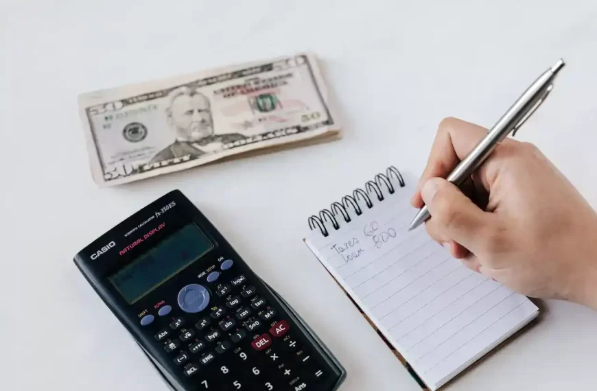 Close up of a persons hand writing a budget in a notepad with calculator and money on the table