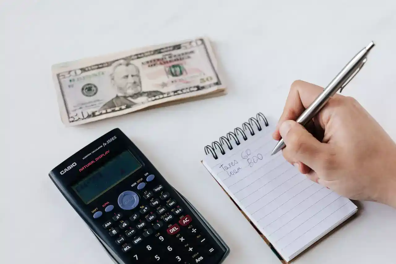 Close up of a persons hand writing a budget in a notepad with calculator and money on the table