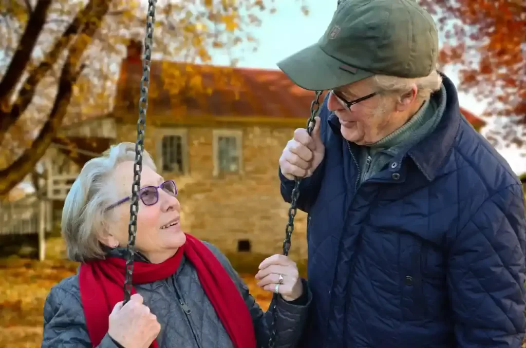 Elderly man standing beside an elderly woman on a swing, smiling at each other