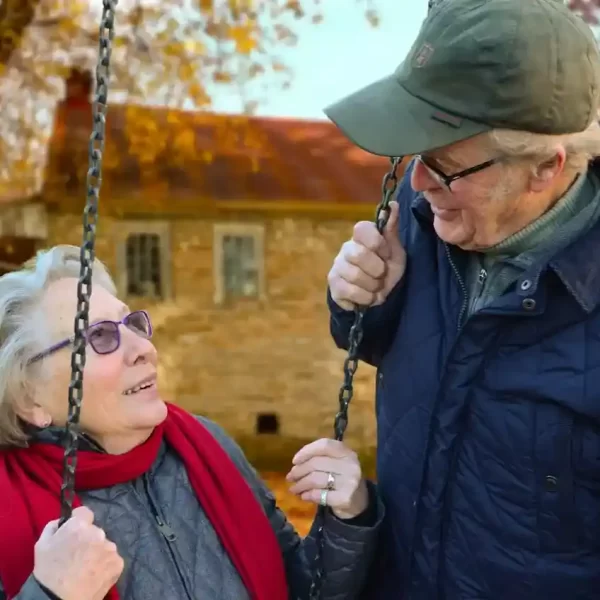 Elderly man standing beside an elderly woman on a swing, smiling at each other