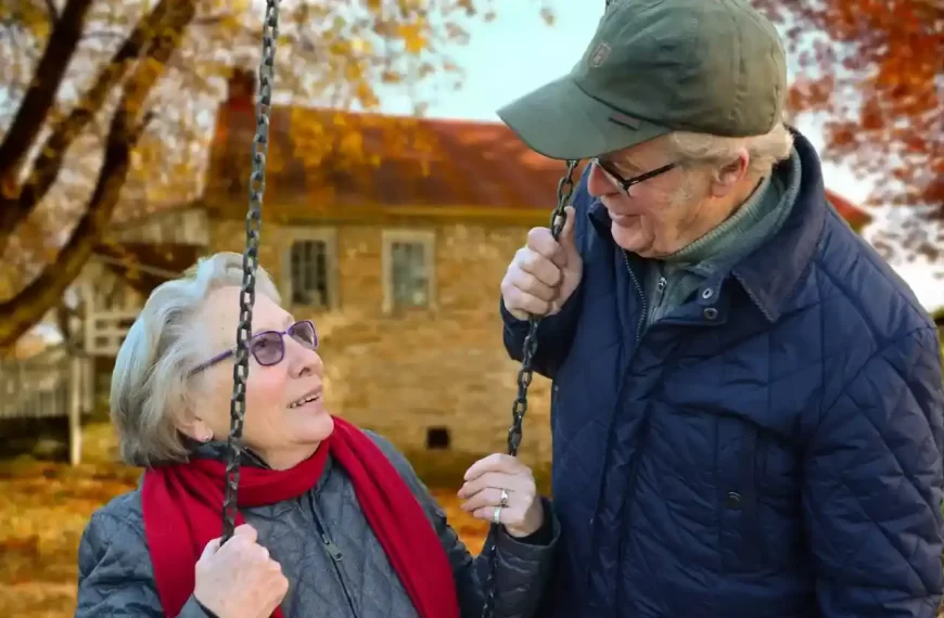 Elderly man standing beside an elderly woman on a swing, smiling at each other