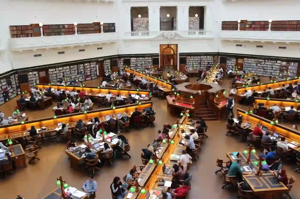 Many people sitting inside a room that looks like a library, looking at computers