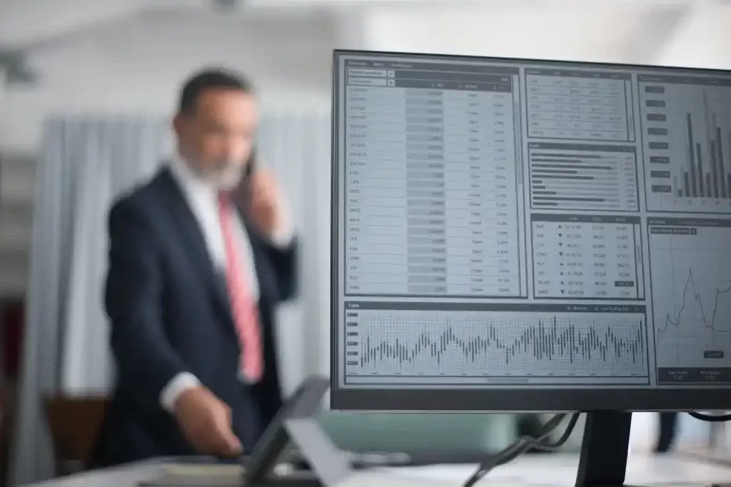 Stock market data displayed on computer with an out of focus man calling someone in the background