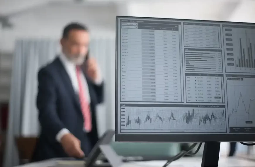 Stock market data displayed on computer with an out of focus man calling someone in the background