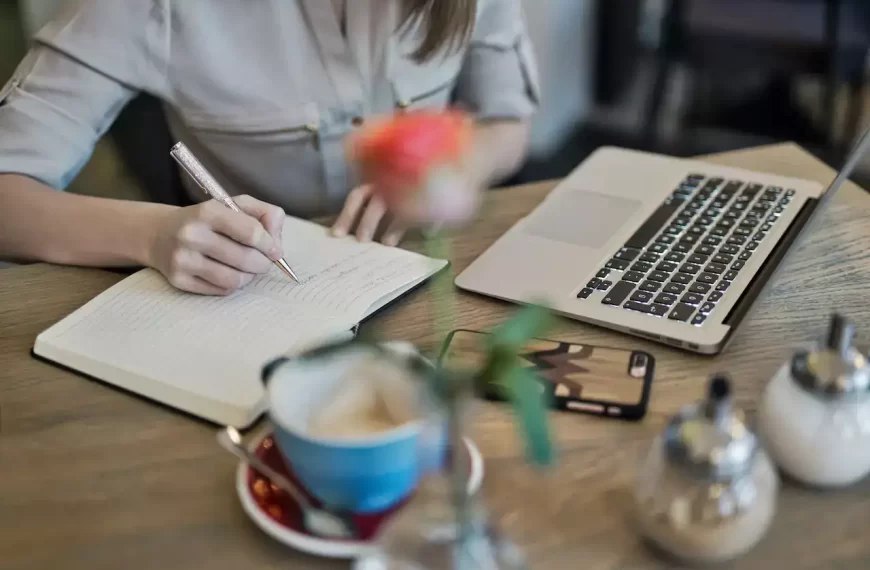 A woman writing in a notebook in front of a laptop