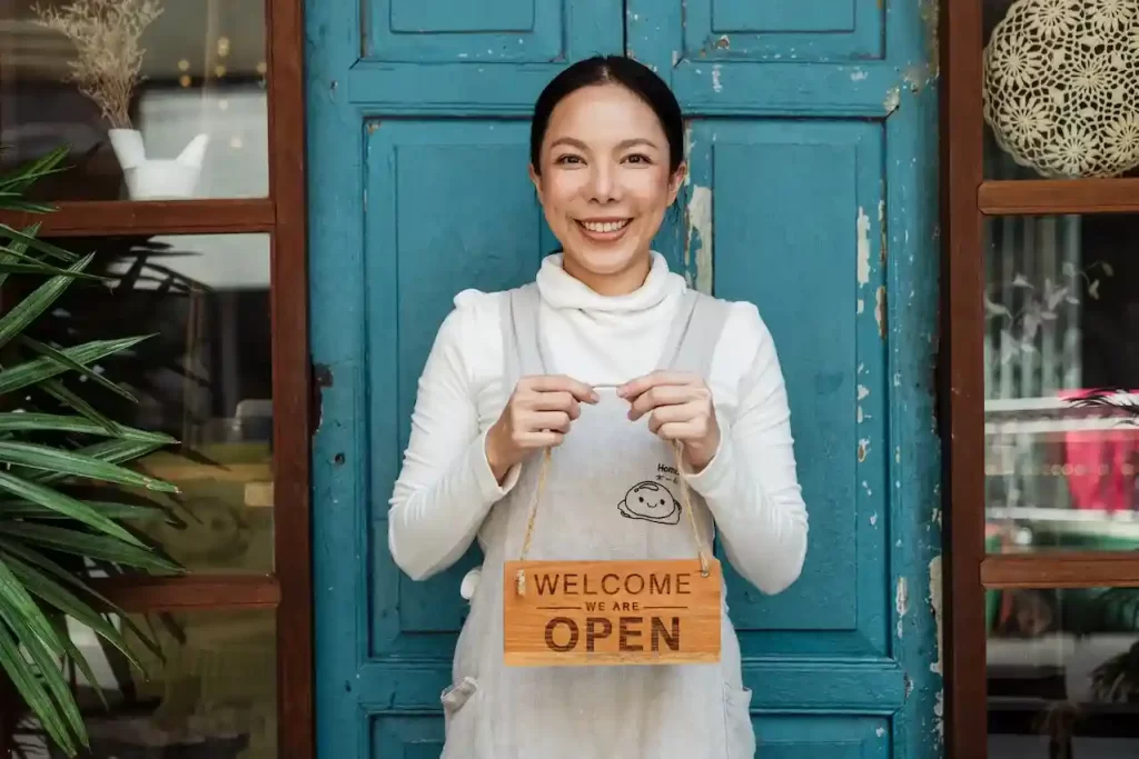 A woman standing in front of a door, smiling with a "Welcome Open" sign in her hands